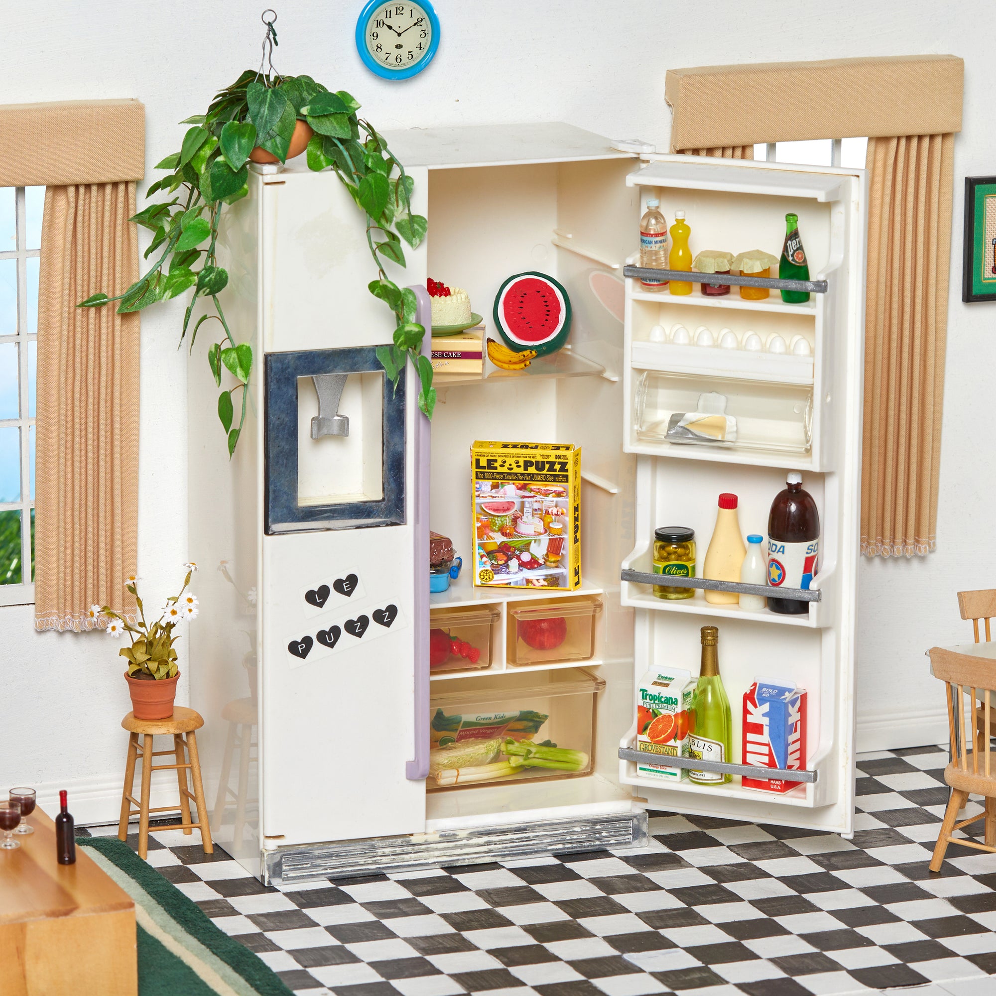 A miniature white toy refrigerator with its door open, filled with colorful pretend food items, set in a dollhouse-style room with a checkered floor and curtains.