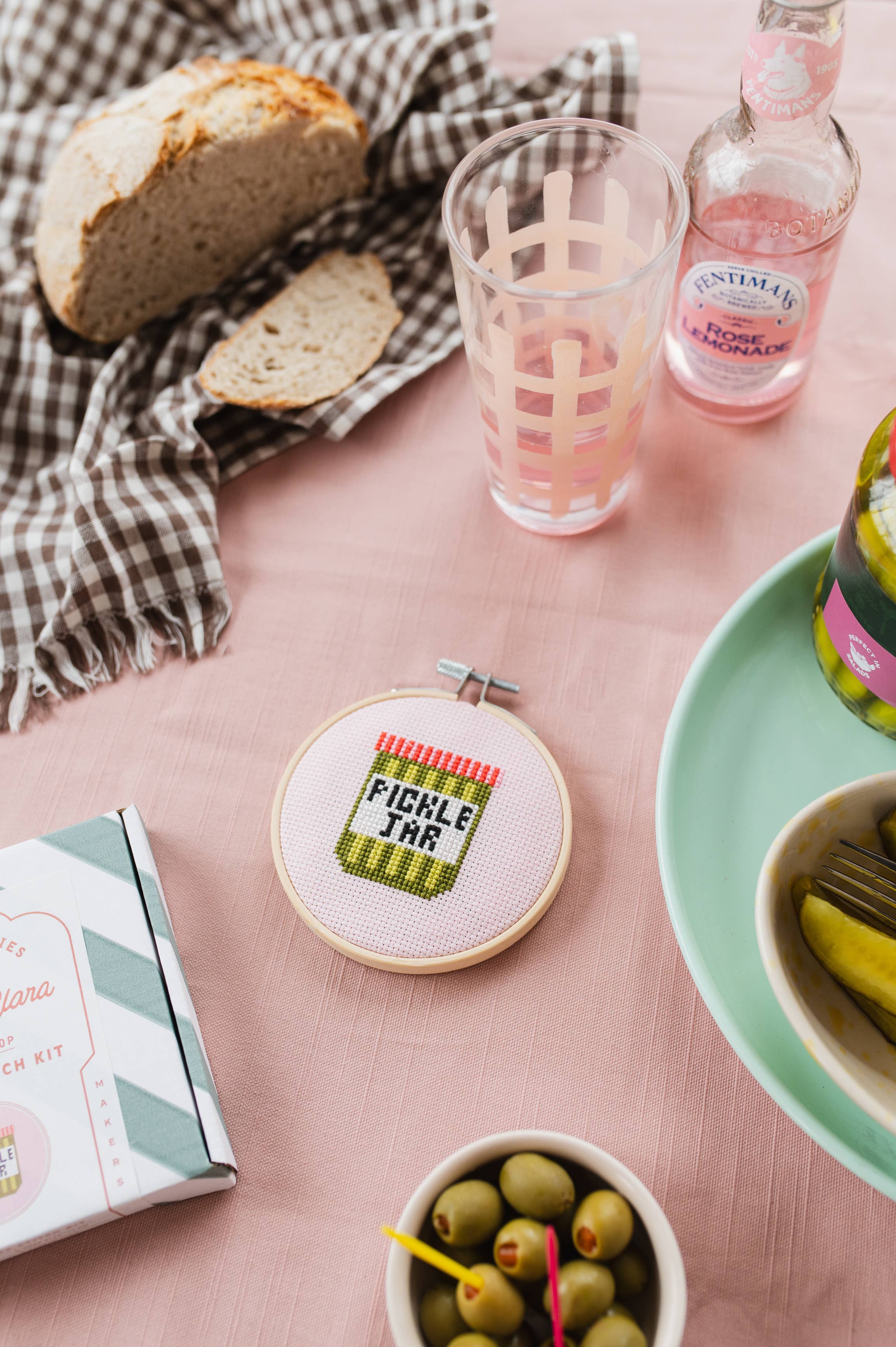 Table setting with bread, glass of pink lemonade, bottle of pink lemonade, and pickle jar cross-stitch kit.