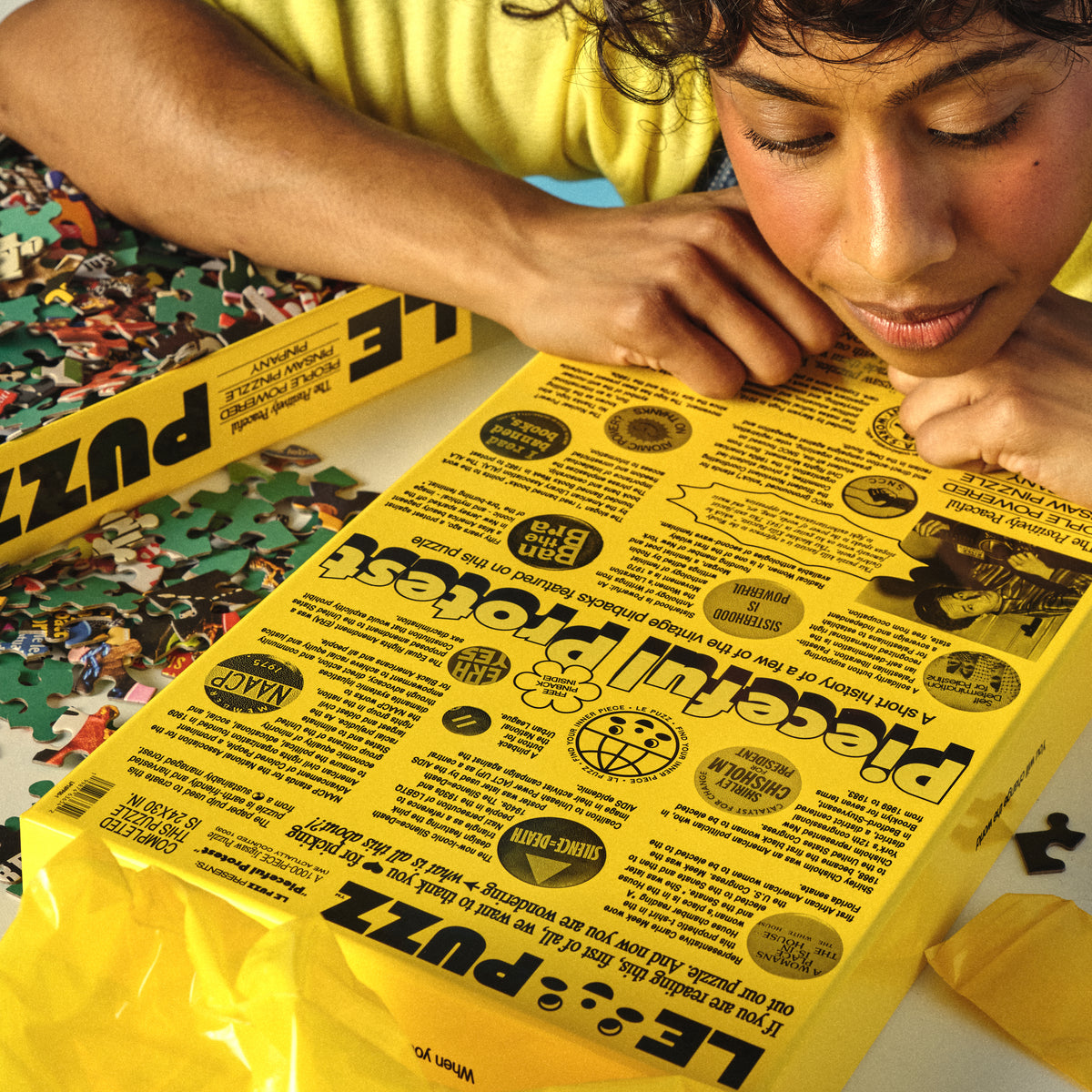 Person looking at a yellow puzzle box with text and graphics on a table with a partially completed puzzle.