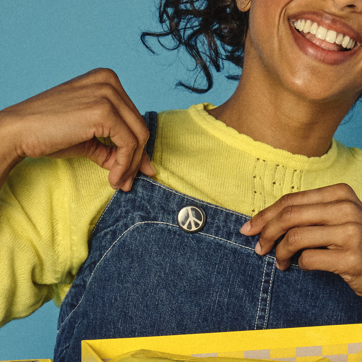 Person wearing a yellow sweater and blue denim overalls with a peace sign button, against a blue background.