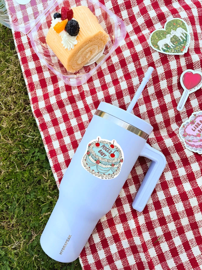 Light blue tumbler with a cake design on a red and white checkered tablecloth with a dessert in the background.