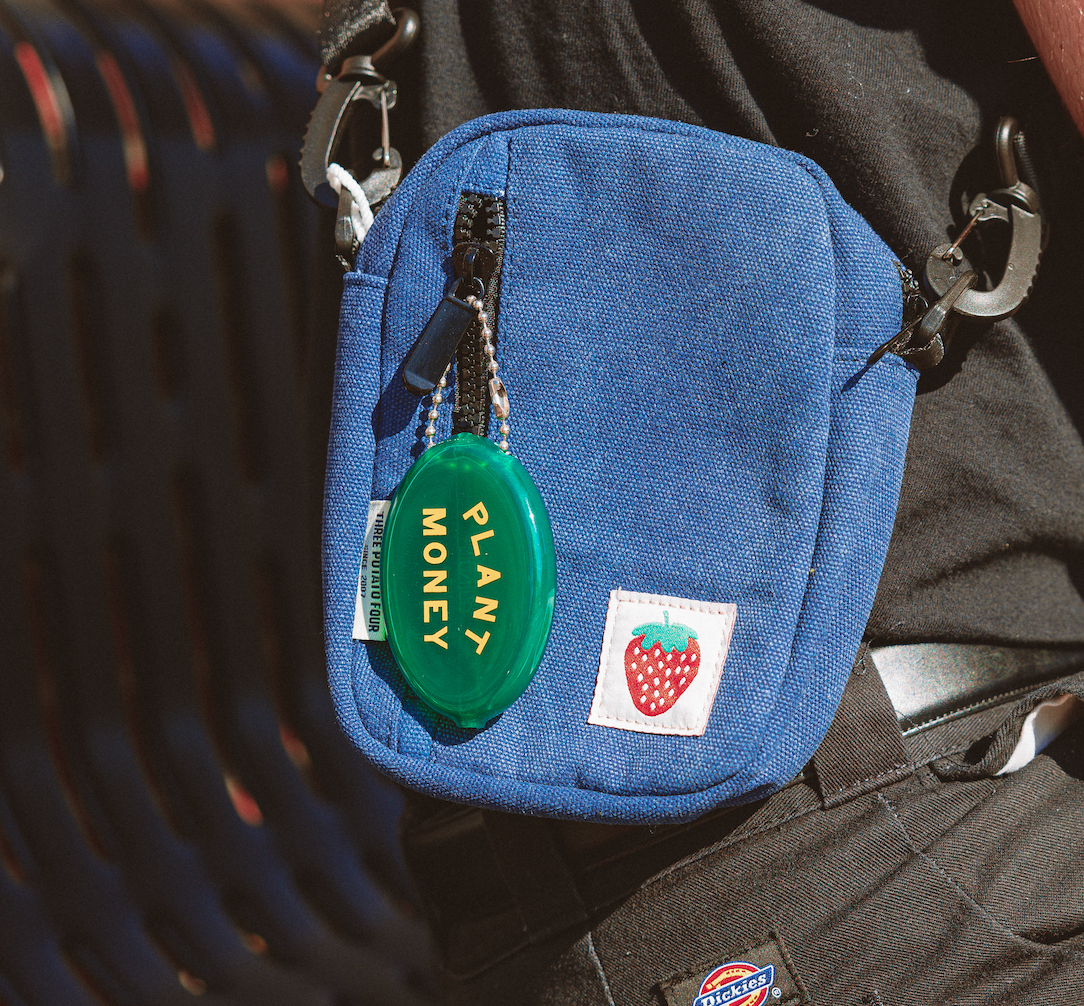 Blue pouch with a green 'Plant Money' tag and a strawberry logo on a person's pocket.
