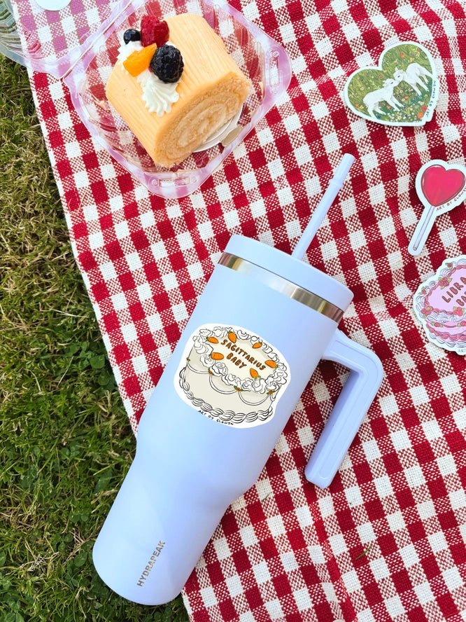 Light blue tumbler with a cake design on a red and white checkered tablecloth with a dessert and decorative elements.
