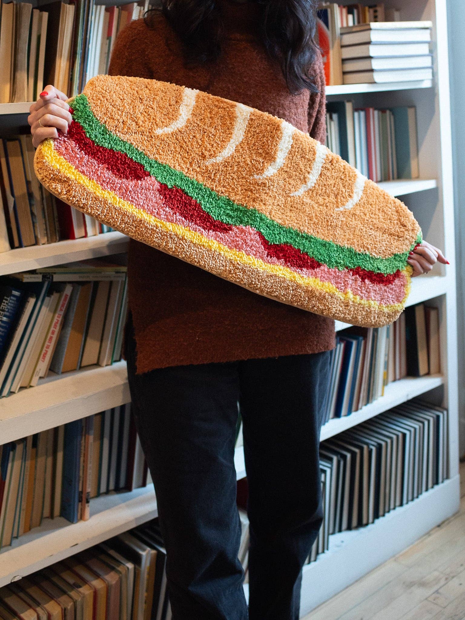 Person holding a large, colorful rug shaped like a sandwich in a room with bookshelves.