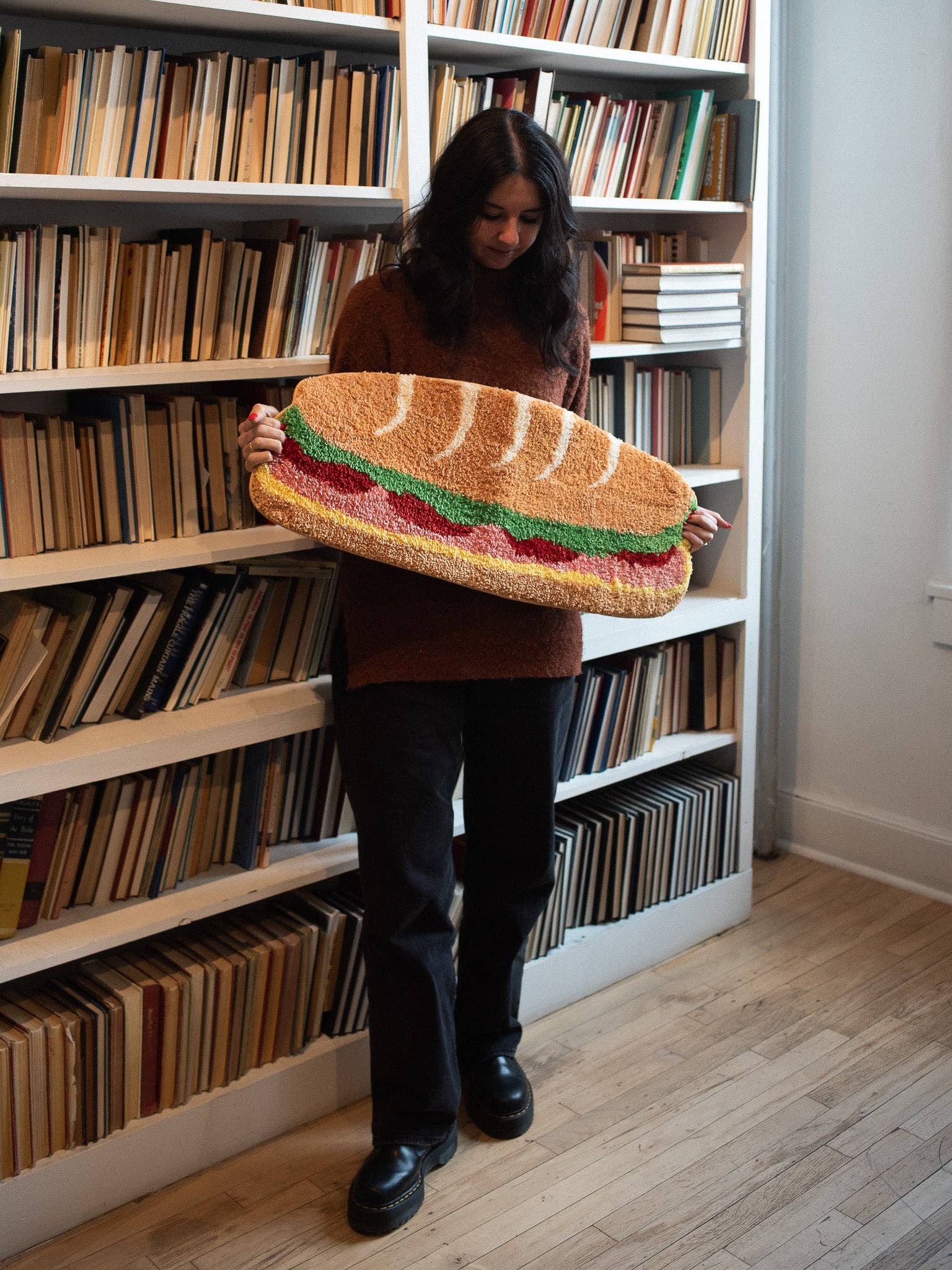 Person holding a large sandwich-shaped pillow in front of bookshelves
