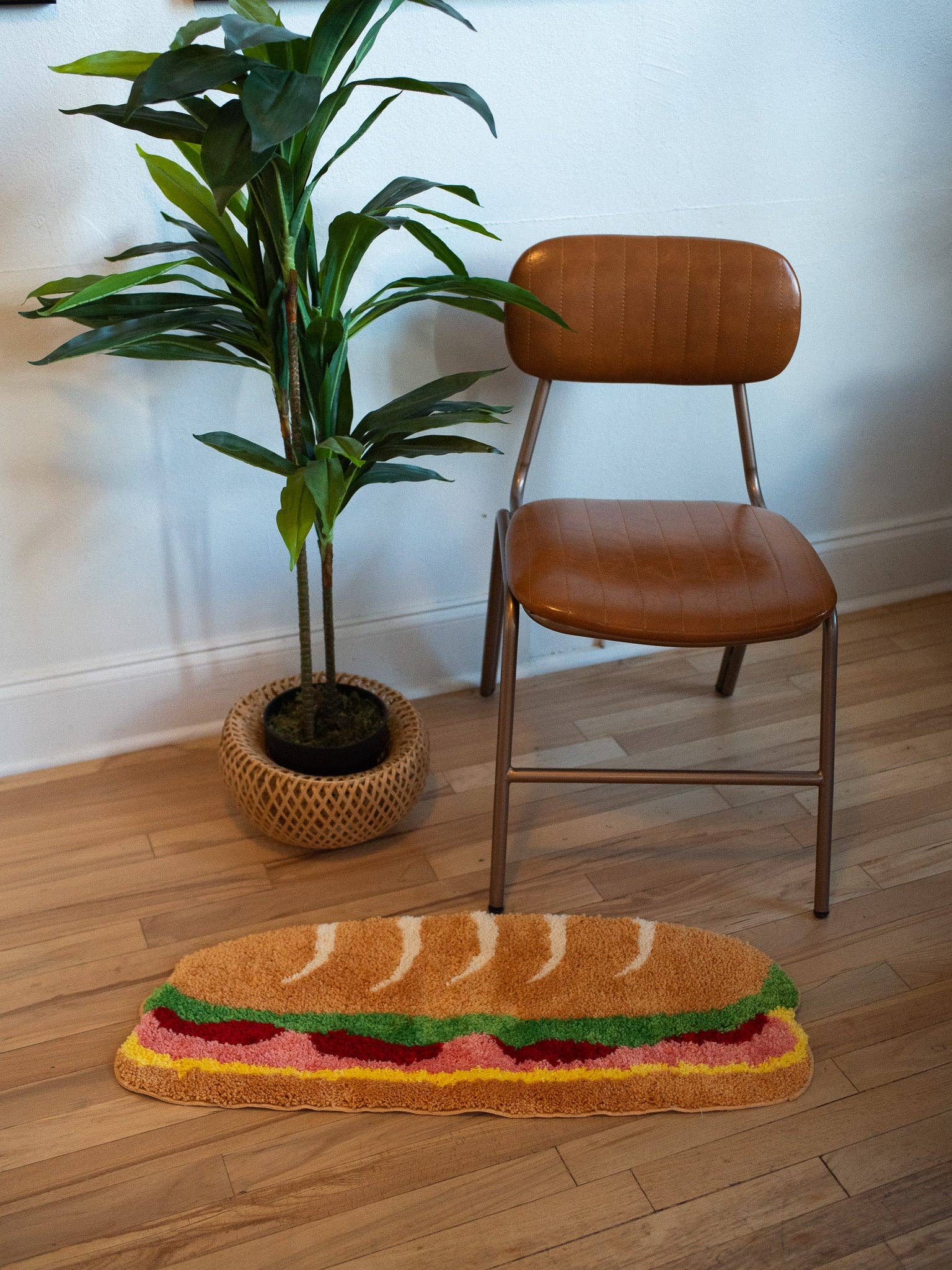 Brown chair next to a plant and a rug shaped like a sandwich on a wooden floor.