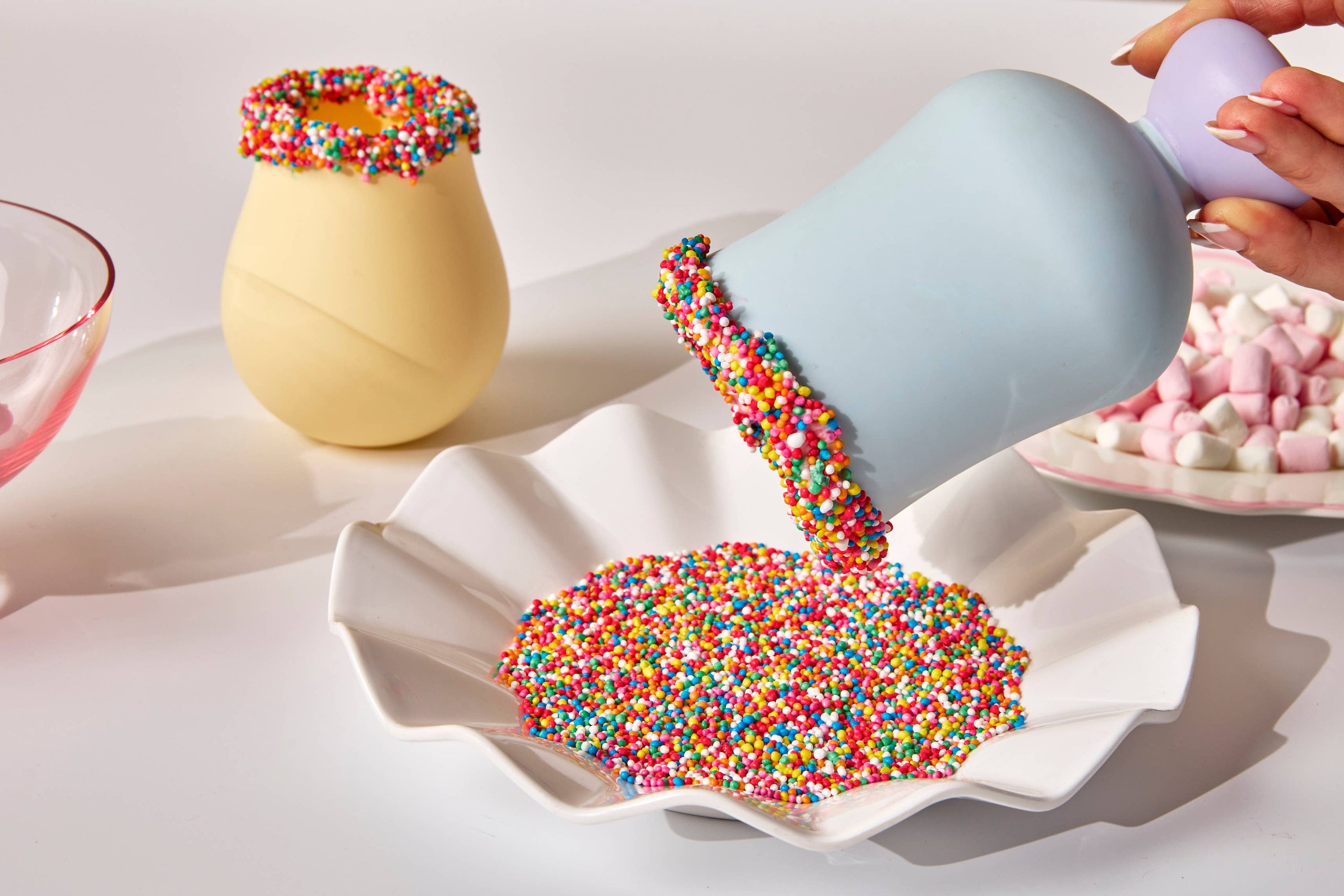 Colorful sprinkles being poured from a blue cup into a white dish on a light surface.