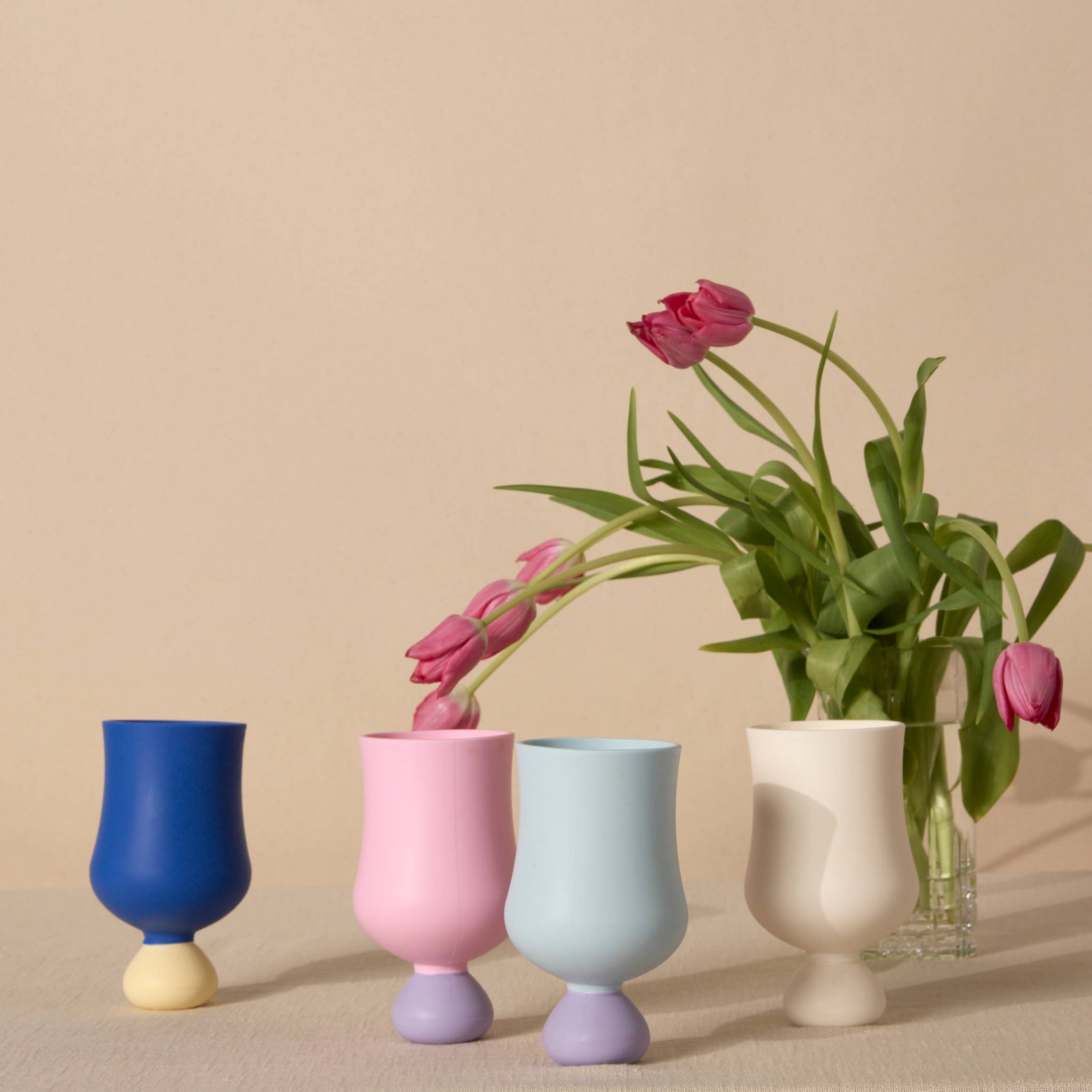 Set of four colorful silicone goblet cups with a vase containing pink tulips on a beige background