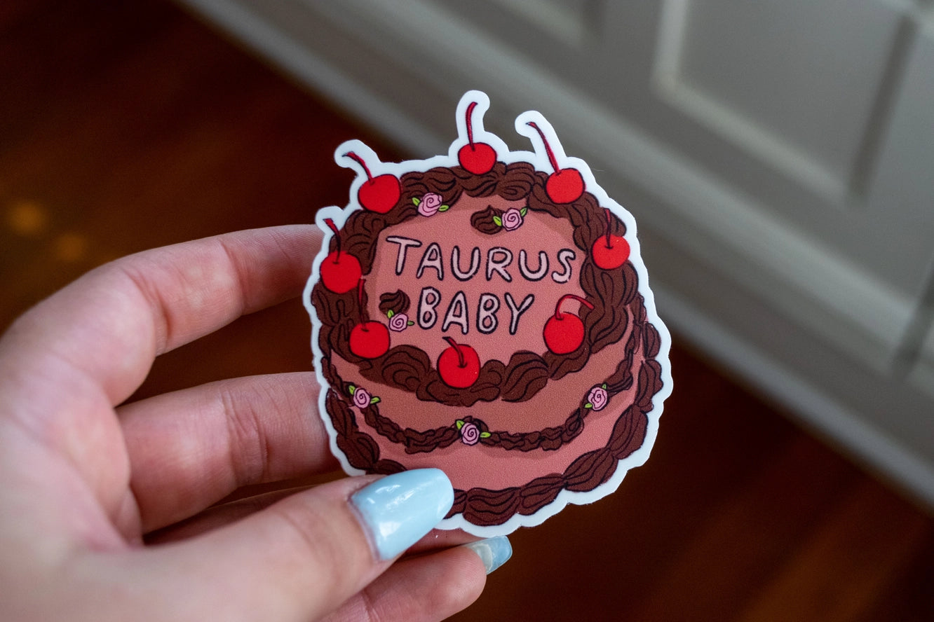 Sticker of a cake with 'Taurus Baby' text held by a hand.