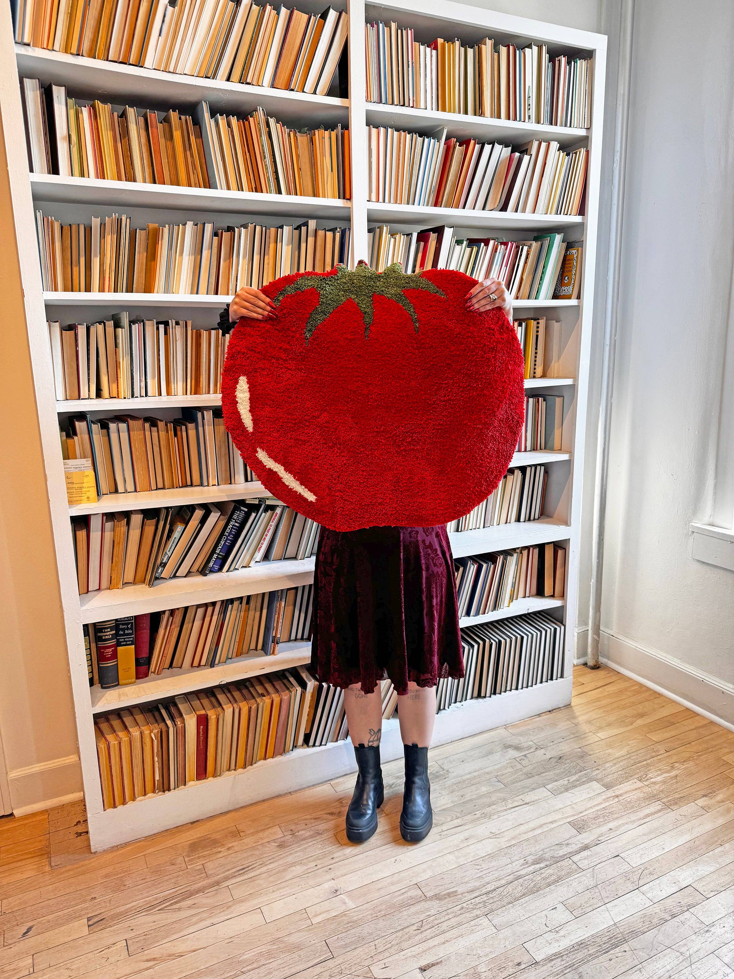 Person holding a large red tomato plush toy in front of a bookshelf