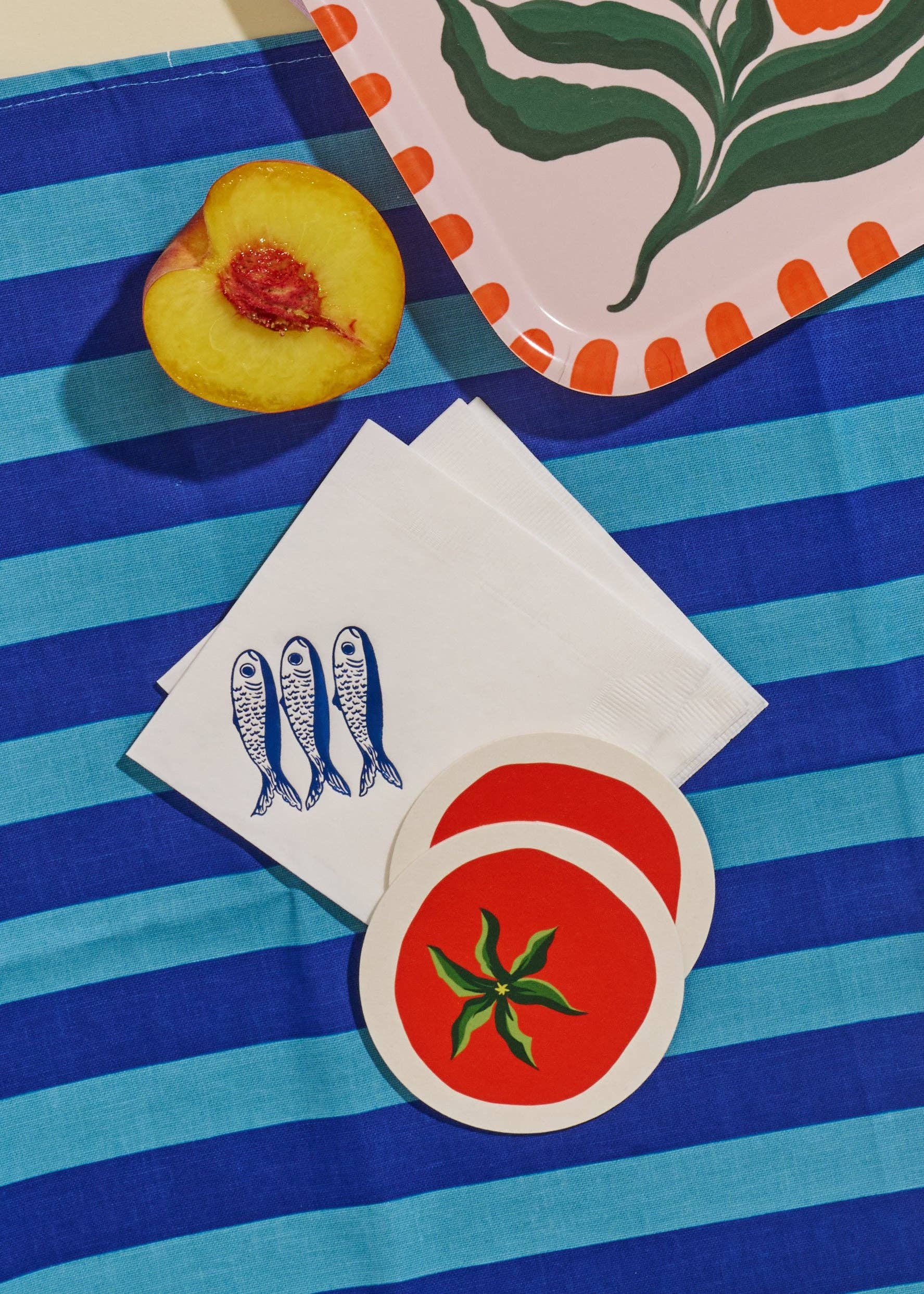 Stack of white napkins with blue fish design, red coaster with green leaf design on a blue and white striped surface.