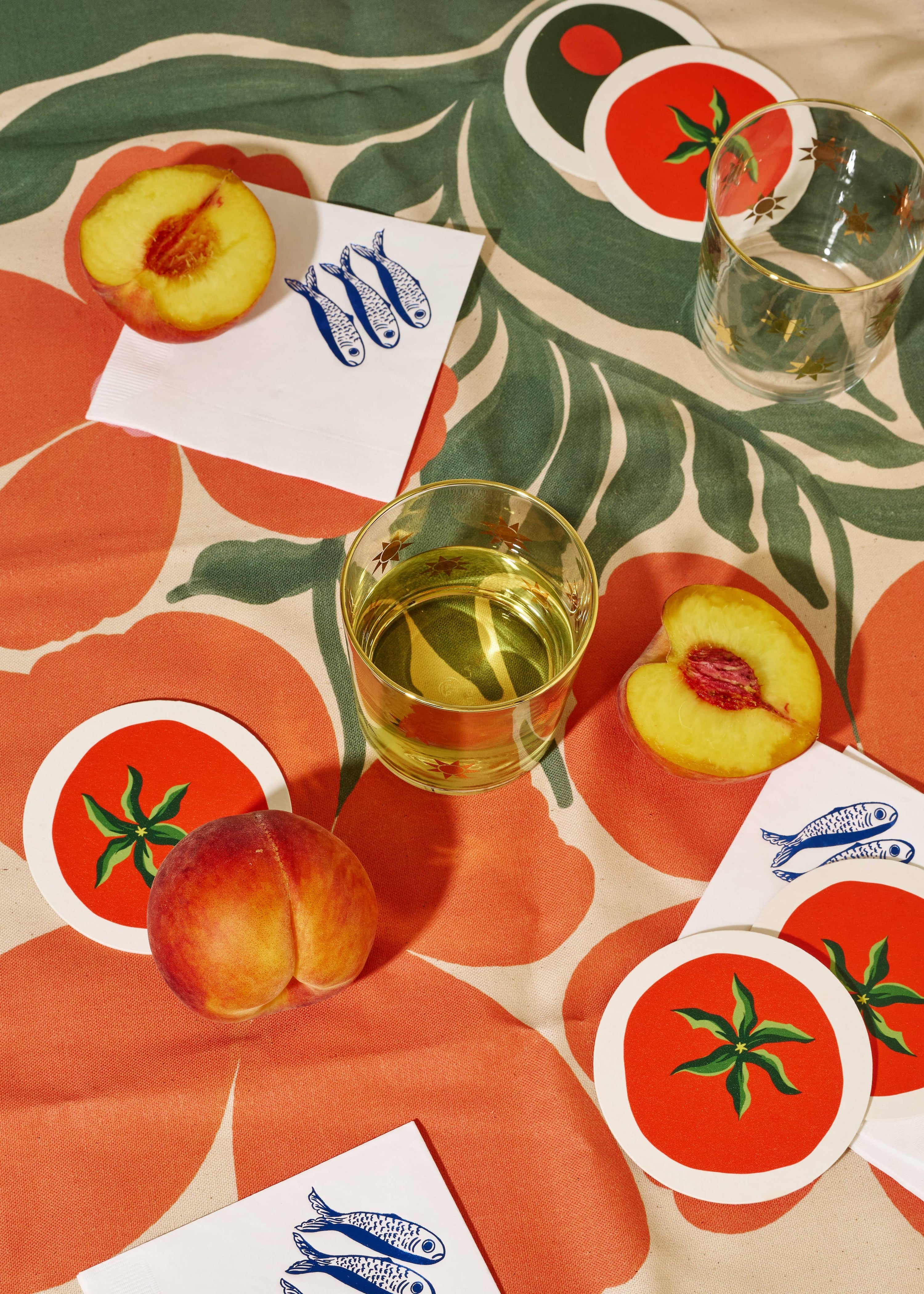Table setting with peaches, a glass of white wine, and coasters on a patterned tablecloth.