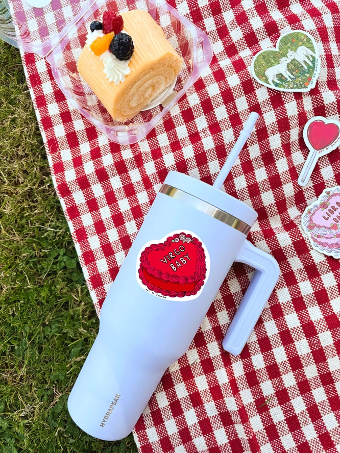 White tumbler with a red heart design on a red and white checkered blanket with a cake and decorative elements.